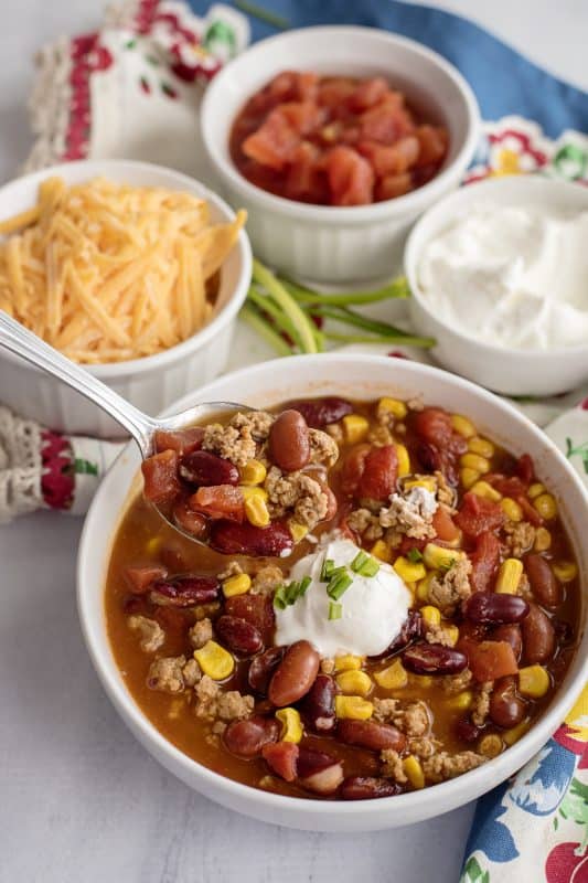 Picture of Taco Soup served in a round white bowl with a dollop of sour cream. Behind the bowl are little bowls filled with toppings for the soup - shredded cheddar cheese, diced tomatoes and sour cream. 