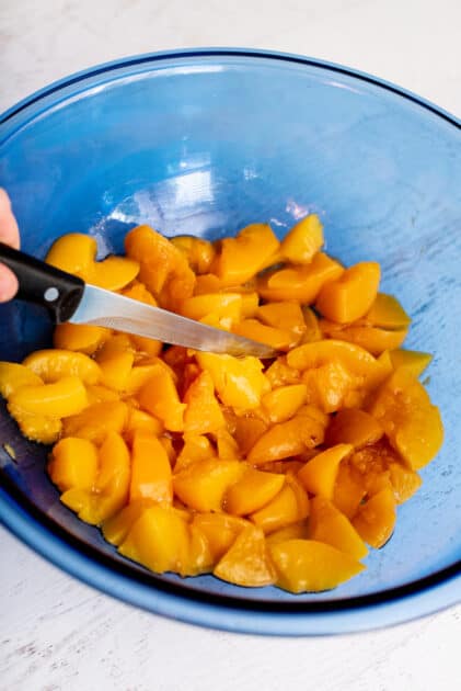 Chopped peaches in a glass blue mixing bowl with a knife.