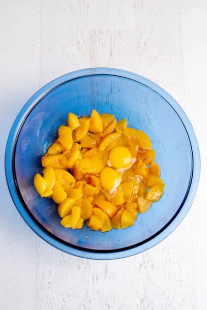 Two eggs in a glass blue mixing bowl with chopped peaches.