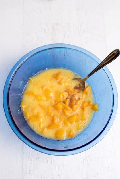 Adding vanilla to a glass blue mixing bowl of chopped peaches and whisked eggs.