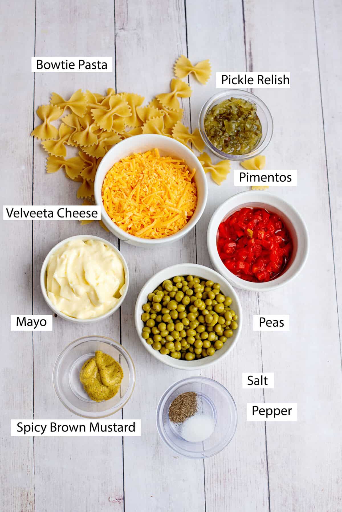 Ingredients for Velveeta bowtie pasta, featuring Velveeta cheese, bowtie pasta, mayo, English peas, spicy brown mustard, jar of pimentos, sweet pickle relish, salt, and pepper.
