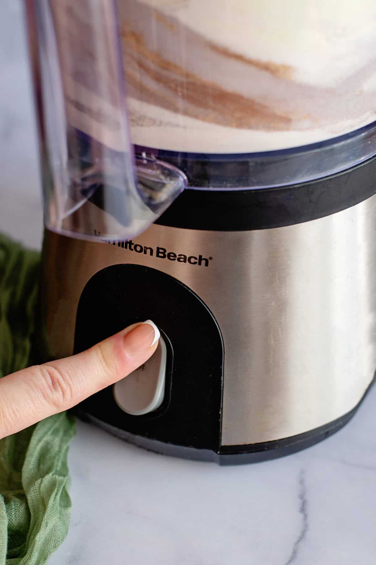 A finger pressing the button of a food processor with swirls of vanilla chai powder.