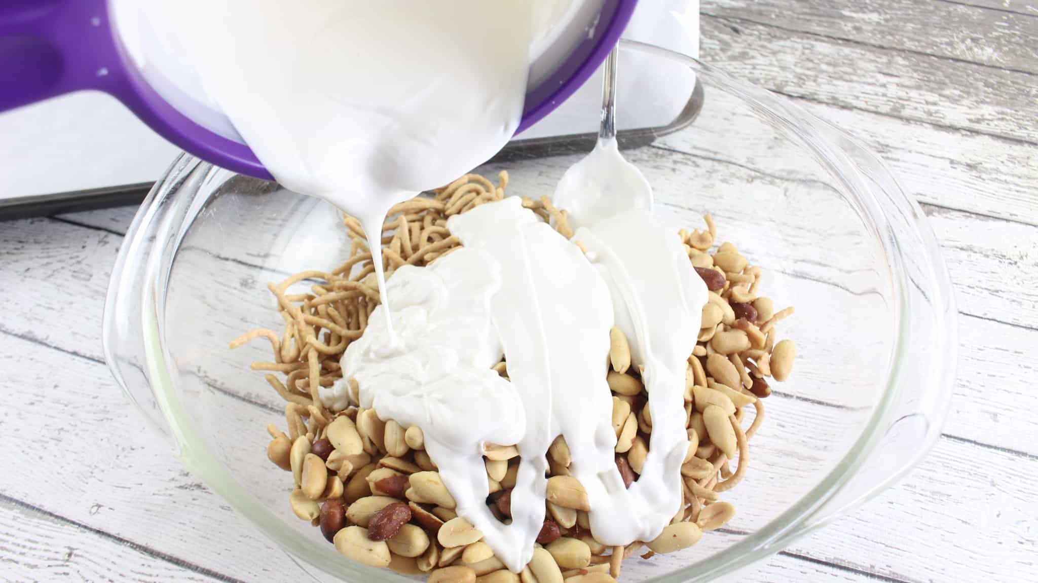 Pouring melted white almond bark over a mixing bowl of chow mein noodles and cocktail peanuts.