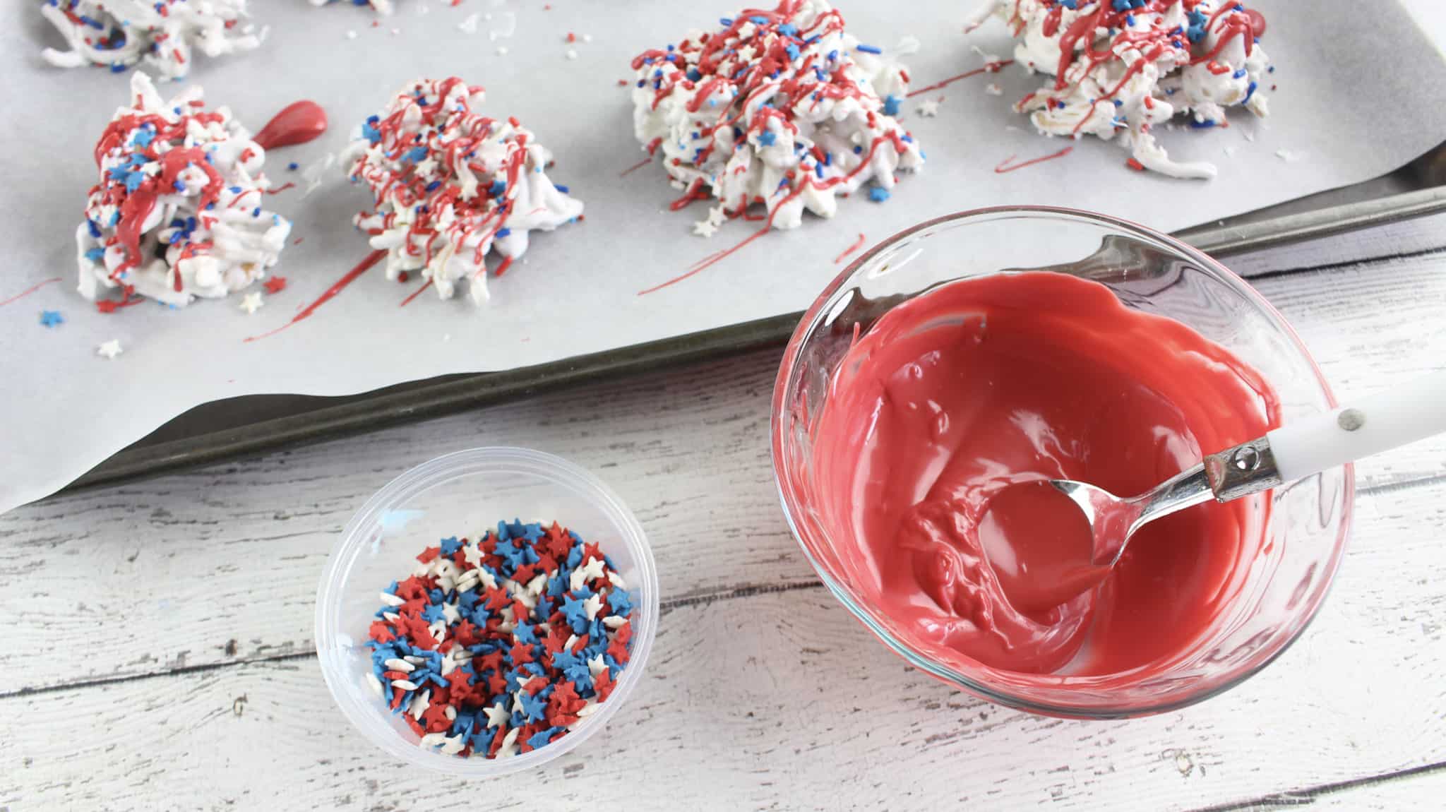 Melted red chocolate in a small glass mixing bowl beside a small cup of sprinkles and a baking sheet of old glory haystack candy.