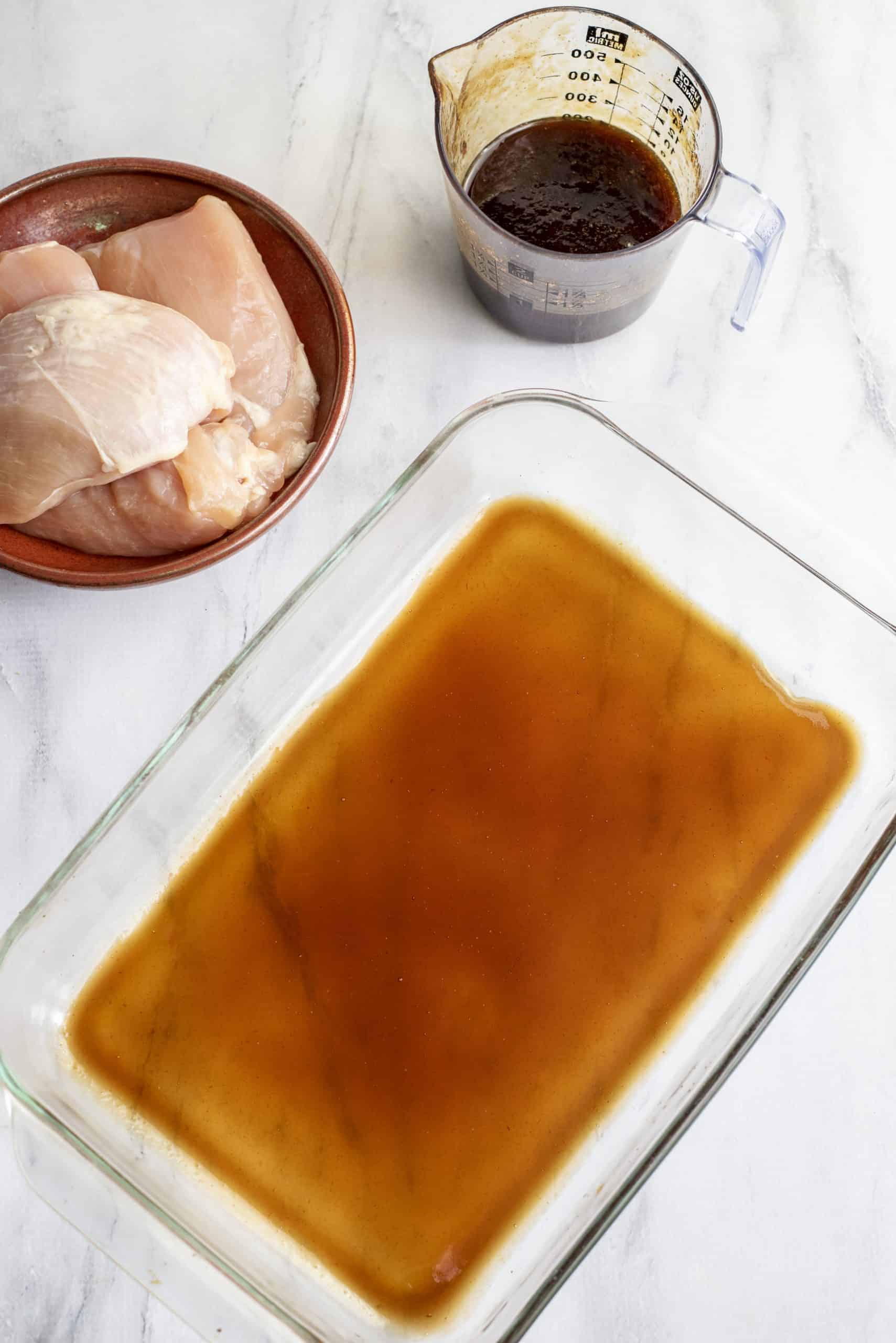 A glass baking dish with BBQ sauce covering the bottom beside raw chicken breast and a measuring cup of BBQ sauce.