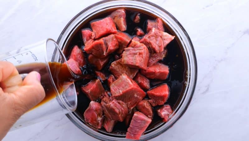 Pouring beef kabob marinade over steak chunks in bowl.