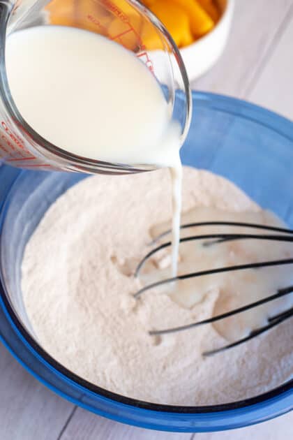 Pouring milk into a glass blue mixing bowl of dry ingredients.