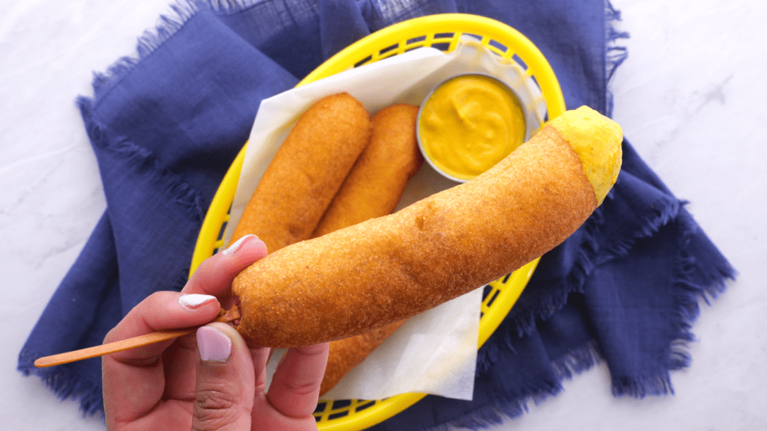 Close-up of corn dog with mustard (with basket of corn dogs in background).