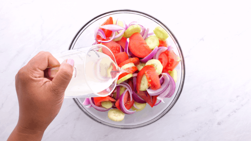 Pouring vinegar mixture over tomato, onion, and cucumber in mixing bowl.