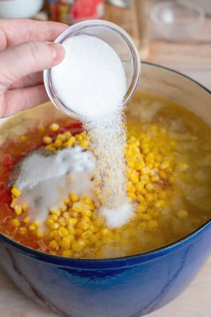 Pouring sugar in a pot of corn, tomatoes, onions, and shredded chicken.