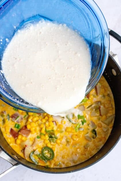 Pouring a mixture of flour and milk into a pot of simmering corn chowder.