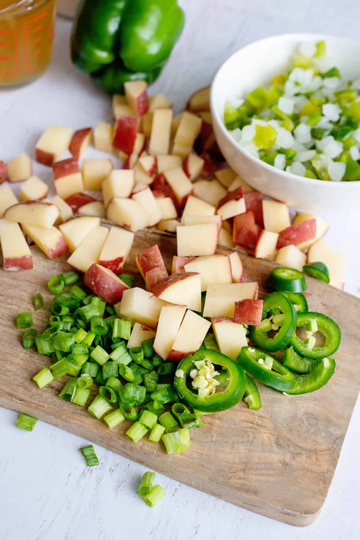 Chopped red potatoes, green bell peppers, and sliced jalapeno on a wooden cutting board.