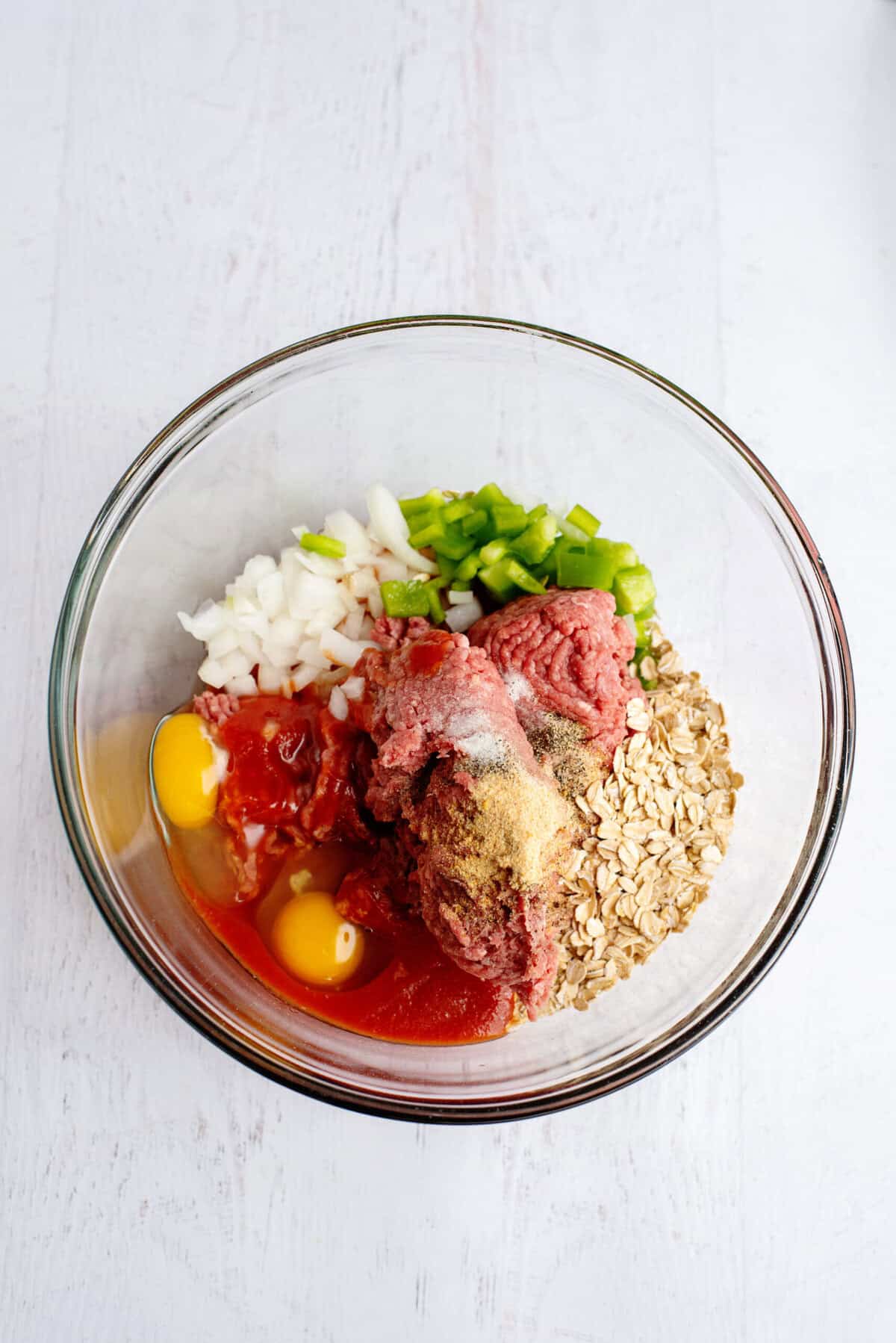 Eggs, tomato sauce, ground beef, quick oats, chopped onion and bell pepper, and spices in a glass mixing bowl before mixing.