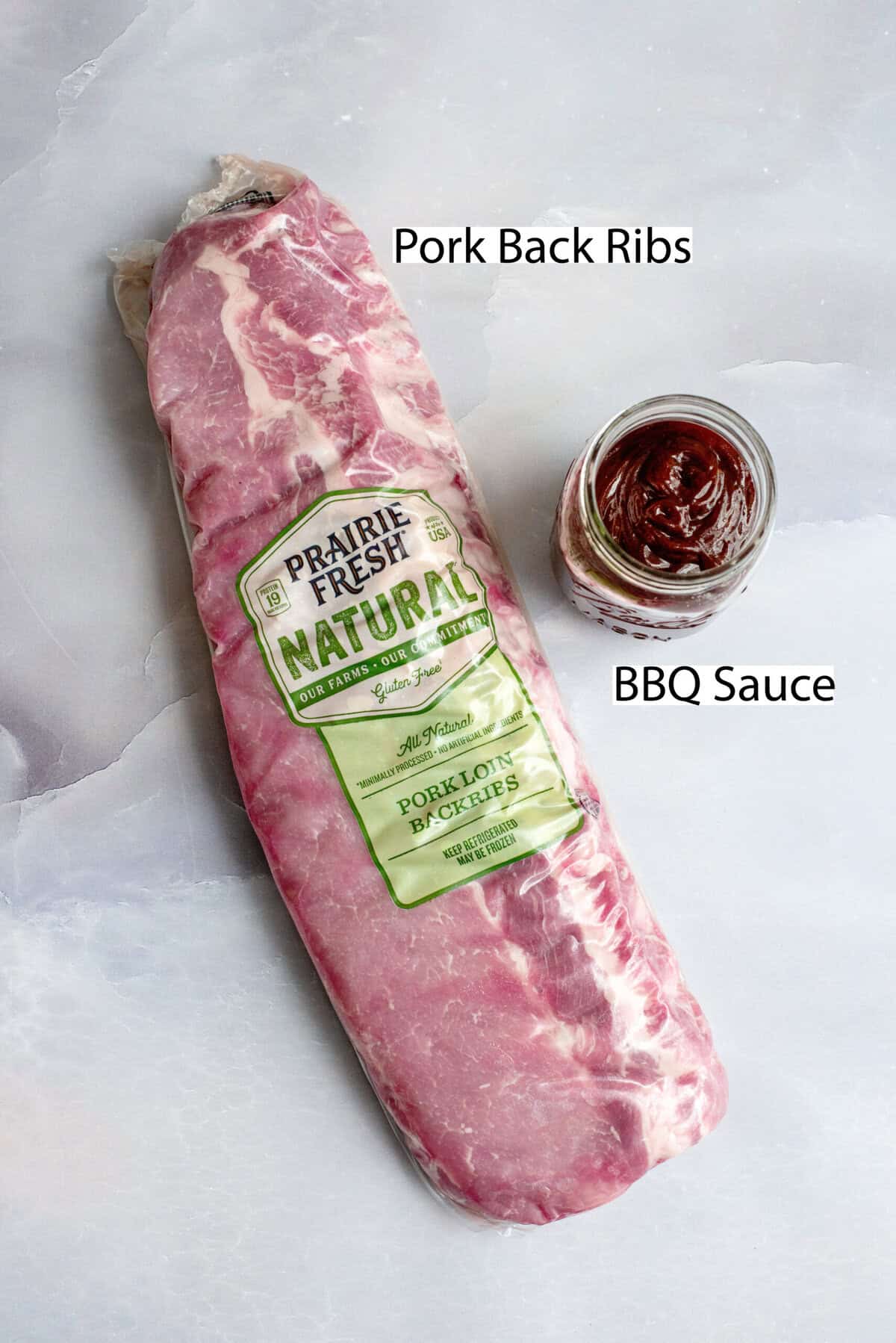 A package of Prairie Fresh Natural pork back ribs placed on a light marble surface next to a jar of thick barbecue sauce. The image is labeled with "Pork Back Ribs" and "BBQ Sauce" to indicate the key ingredients for making oven-baked ribs.
