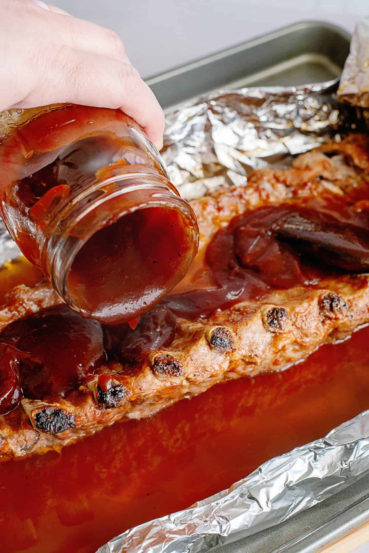 A close-up of barbecue sauce being poured from a jar onto a rack of oven-baked baby back ribs. The ribs are on a foil-lined baking sheet, with a brush nearby for spreading the sauce evenly.