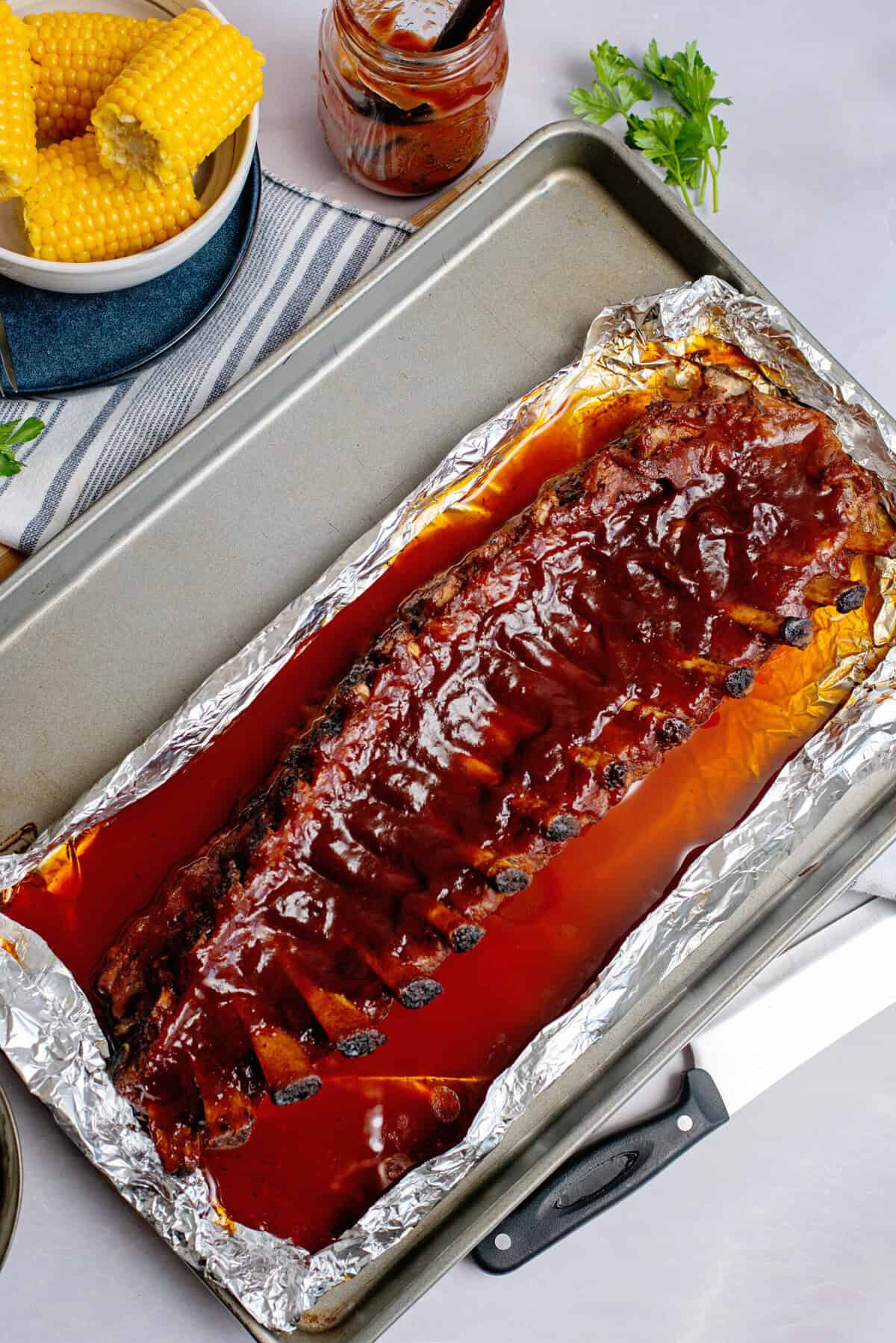 A rack of fully cooked baby back ribs coated in barbecue sauce, resting on a foil-lined baking sheet with juices pooled around it. A jar of BBQ sauce sits in the background on a striped kitchen towel, ready for an extra glaze.