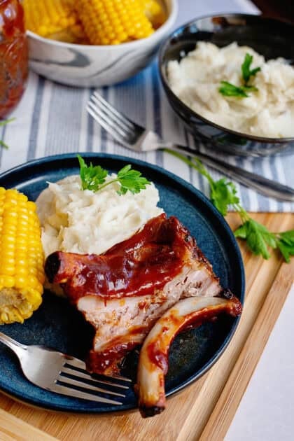 A close-up of a barbecue meal on a dark blue plate, featuring tender, saucy ribs with a deep red glaze, creamy mashed potatoes garnished with a sprig of fresh parsley, and a bright yellow ear of corn on the cob. A silver fork rests on the plate, with a piece of rib meat resting on its tines. The background includes a striped cloth napkin, a glass jar, and additional tableware. The setting is warm and inviting, emphasizing comfort food.