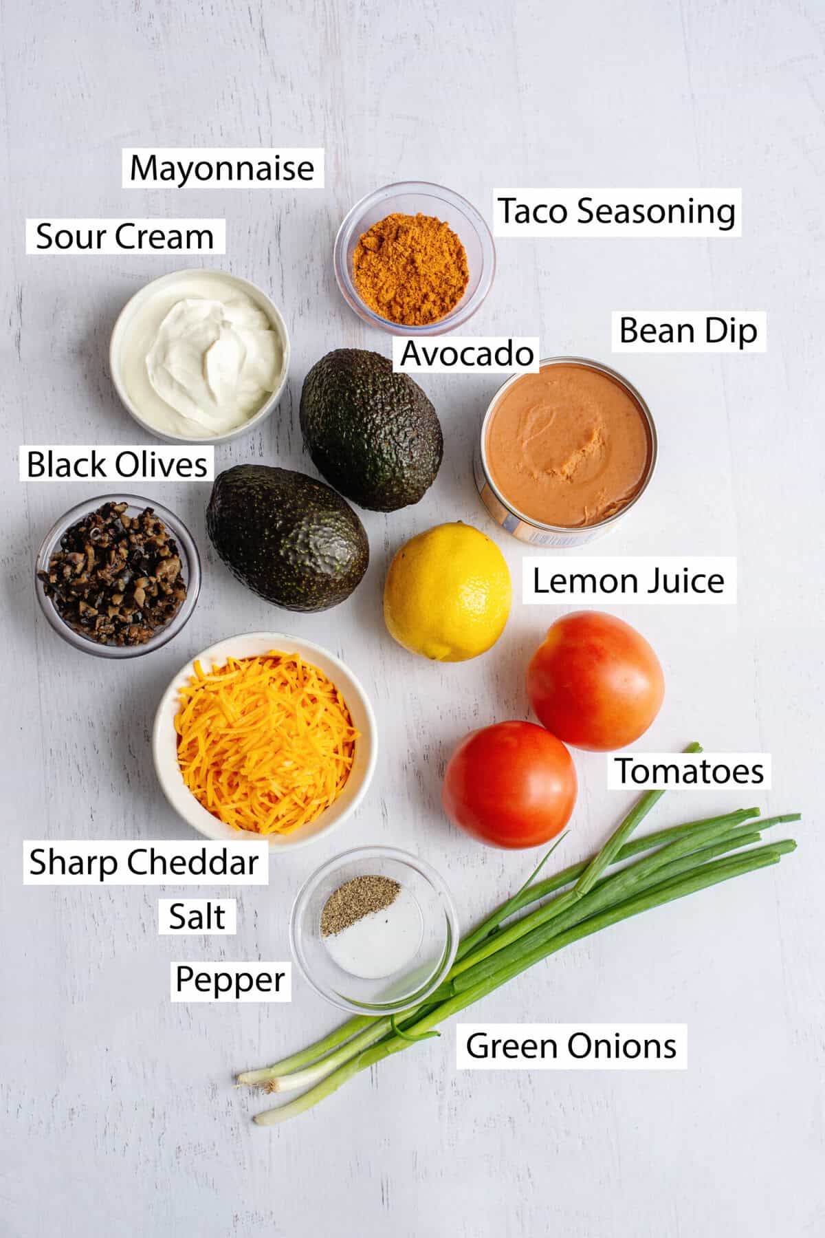 Ingredients for Tex Mex dip, featuring avocados, lemon juice, salt, pepper, sour cream, mayonnaise, taco seasoning, bean dip, green onions, tomatoes, black olives, and shredded cheddar cheese.