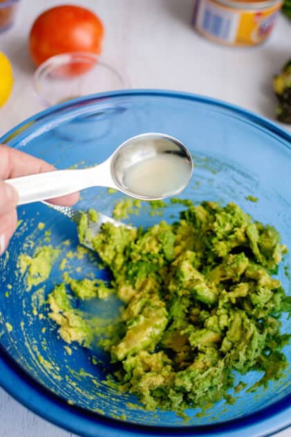 Pouring lemon juice to mashed avocado in a glass blue bowl.