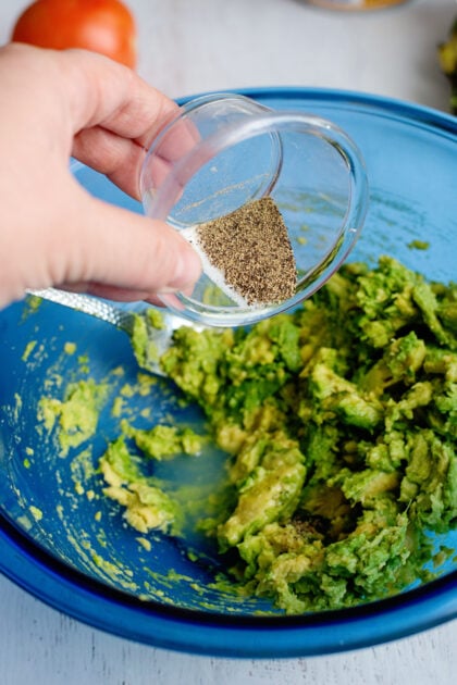 Adding black pepper to mashed avocado in a glass blue bowl.
