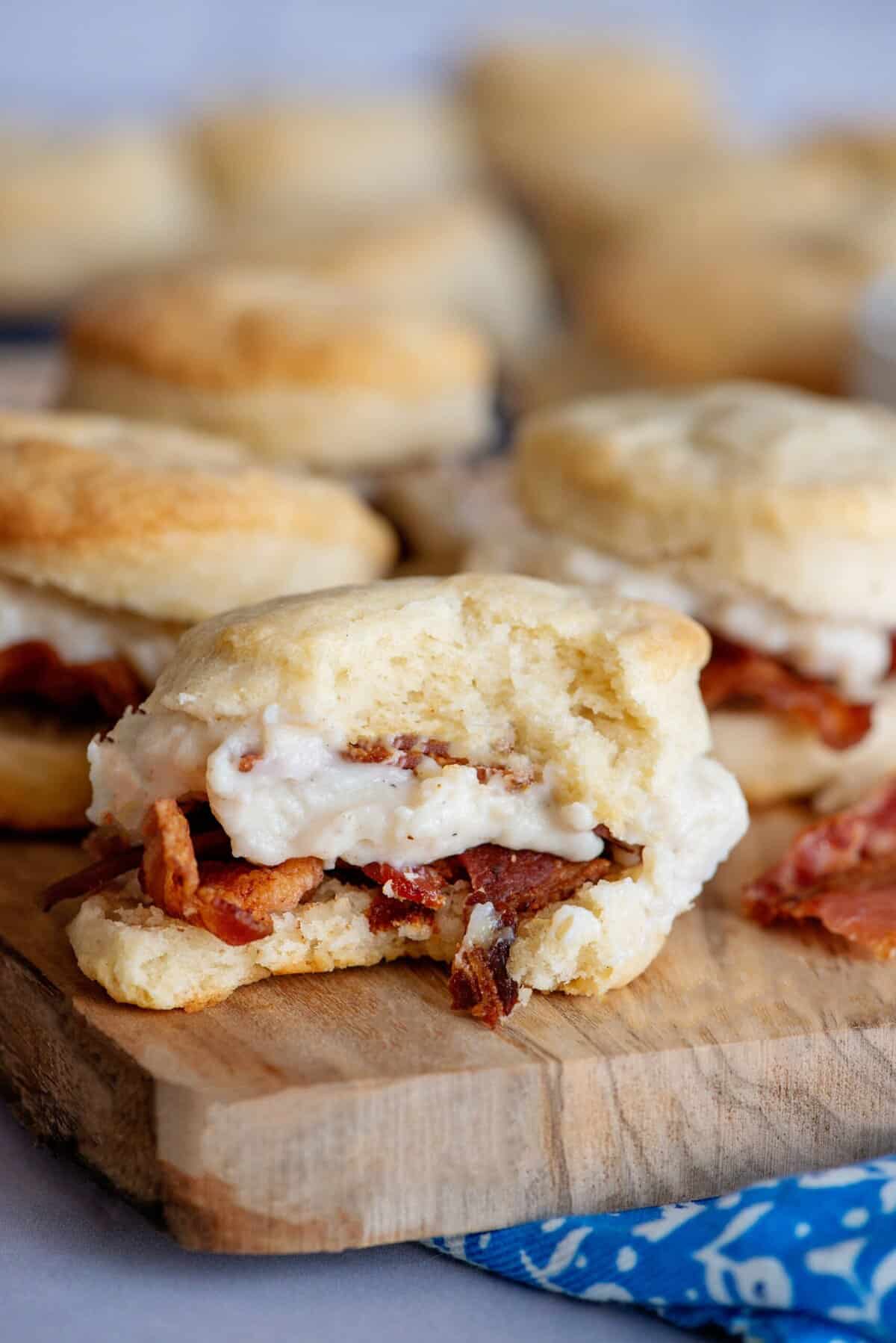 A bitten biscuit with oozing bacon gravy and bacon slices, with rows of biscuits in the foreground.