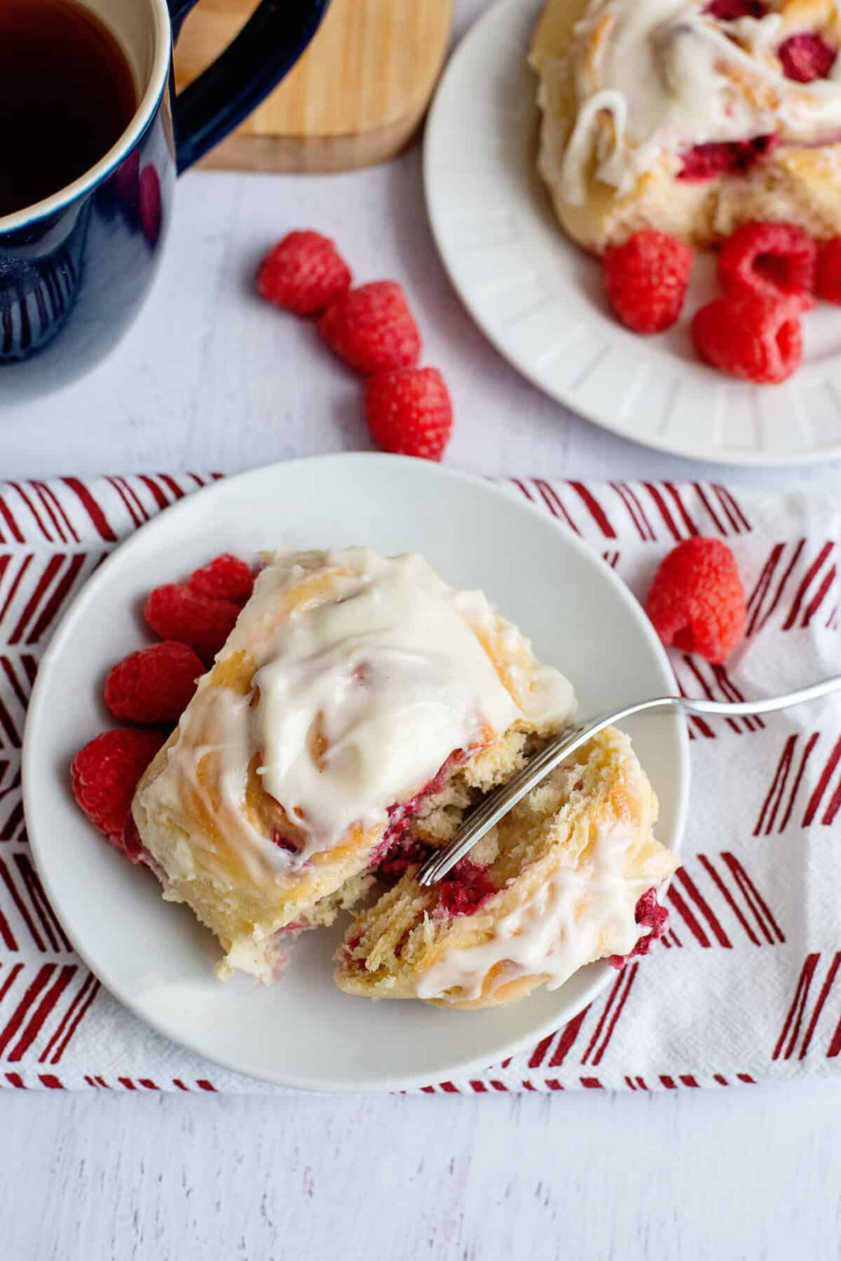 A freshly baked raspberry sweet roll with cream cheese frosting on a white plate, accompanied by fresh raspberries and a cup of tea.