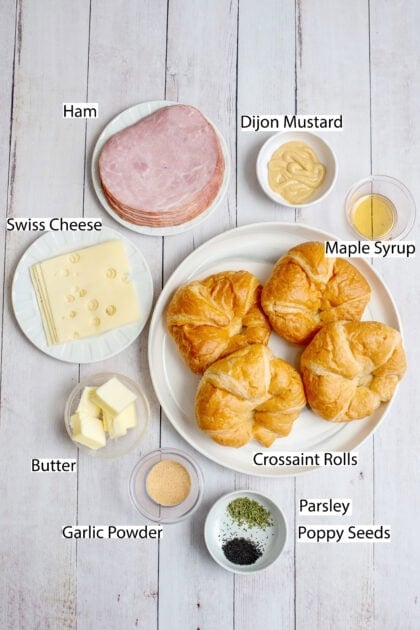 Overhead view of ingredients for ham and cheese croissant sandwiches, including croissant rolls, sliced ham, Swiss cheese, Dijon mustard, maple syrup, butter, garlic powder, parsley, and poppy seeds, arranged on a white wooden surface.