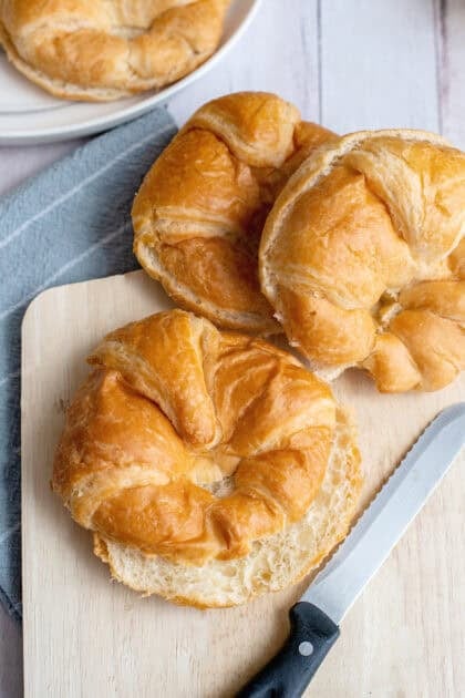 Fresh croissant rolls on a wooden cutting board, with one sliced in half and a serrated knife placed nearby.