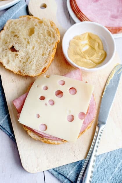 Open croissant sandwich being assembled on a wooden board with layers of ham and Swiss cheese, a bowl of Dijon mustard, a butter knife, and extra ham slices in the background.