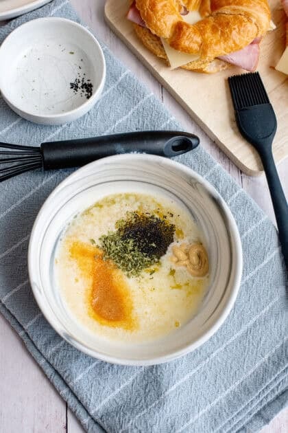 Bowl of melted butter mixed with Dijon mustard, maple syrup, parsley, and poppy seeds, with a pastry brush and prepared croissant sandwiches in the background.