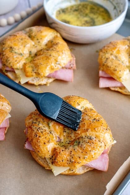 Ham and cheese croissant sandwiches on a baking sheet being brushed with a butter mixture of Dijon mustard, maple syrup, parsley, and poppy seeds before baking.