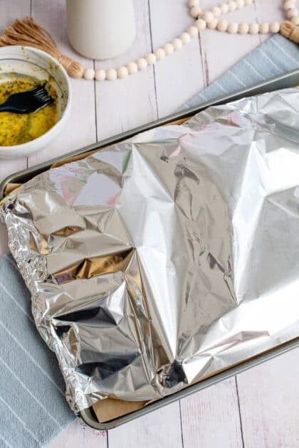 Baking sheet of ham and cheese croissant sandwiches covered with aluminum foil, ready to be baked, with a bowl of butter mixture and brush in the background.