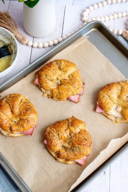 Freshly baked ham and cheese croissant sandwiches on a parchment-lined baking sheet, golden brown and topped with a buttery herb and poppy seed glaze.