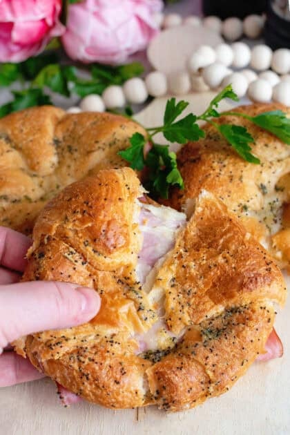 Golden, flaky croissant sandwich filled with melted cheese and slices of ham, topped with poppy seeds and herbs, being pulled apart to reveal its gooey, cheesy center.