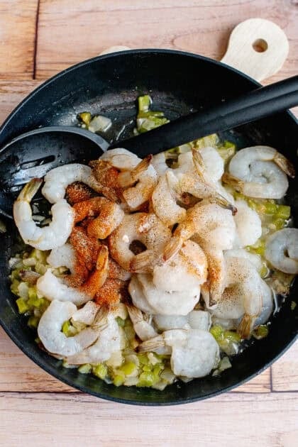 Adding raw shrimp and seasonings to a skillet of simmering vegetables.
