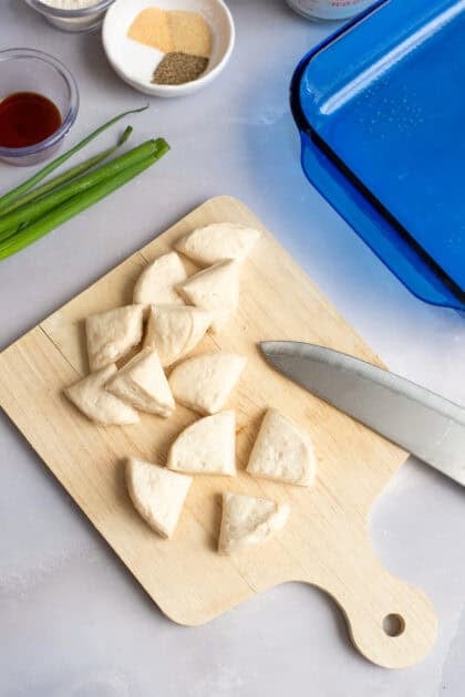 Flaky refrigerated biscuit dough being cut into quarters on a wooden cutting board with a knife.