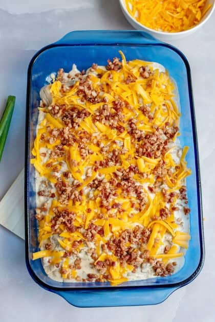 Flaky biscuit dough pieces scattered in a baking dish topped with sausage gravy, bacon bits, and cheddar cheese.