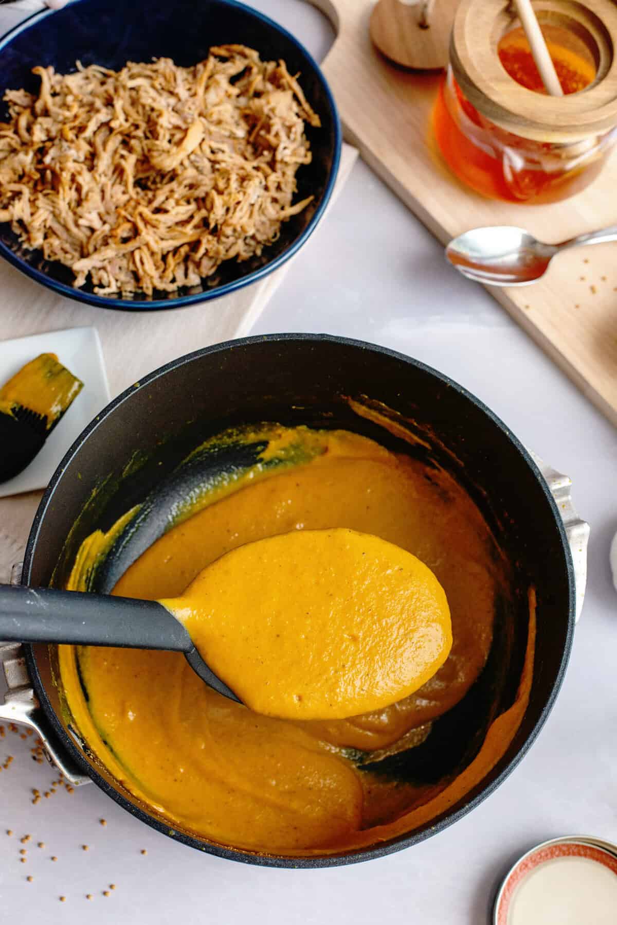 A glossy spoonful of mustard-based Carolina Gold BBQ sauce suspended above a steaming saucepan, ready to be tasted.