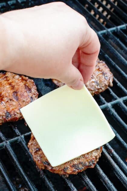 Person placing a slice of cheese on burger patty while it's cooking on the grill