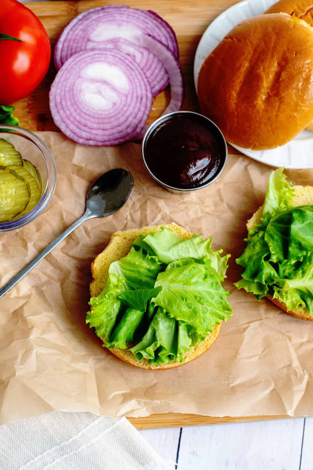 Bottom burger bun with lettuce next to other toppings