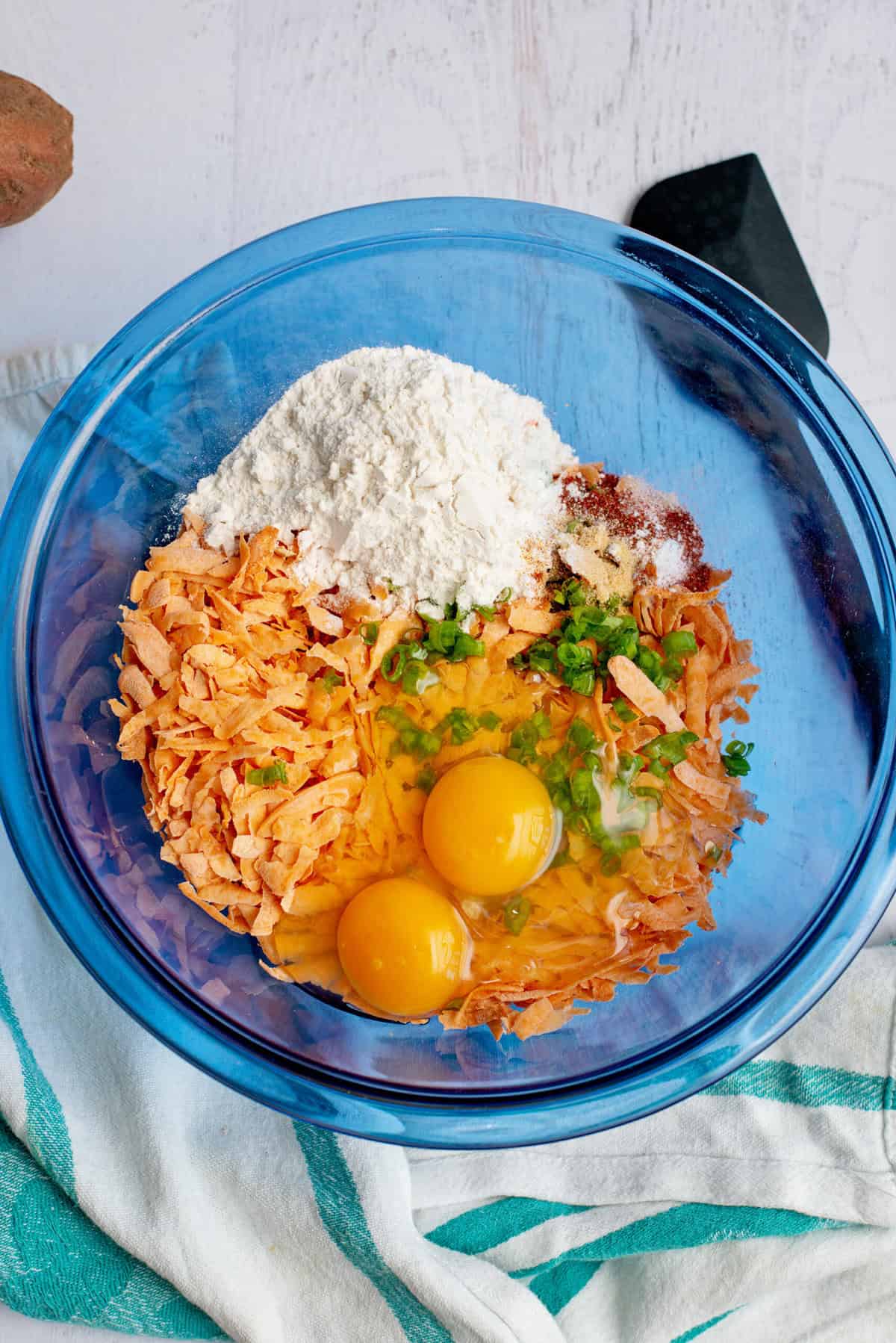 A mixing bowl with grated sweet potatoes, eggs, flour, baking powder, garlic powder, paprika, salt, pepper