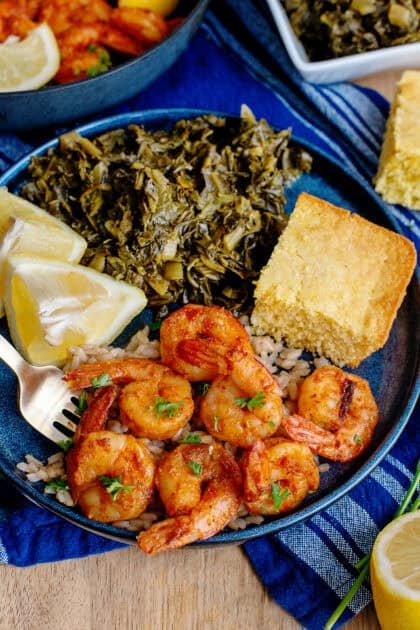 A plate of Cajun shrimp with a side of bread, lemon wedges, and greens