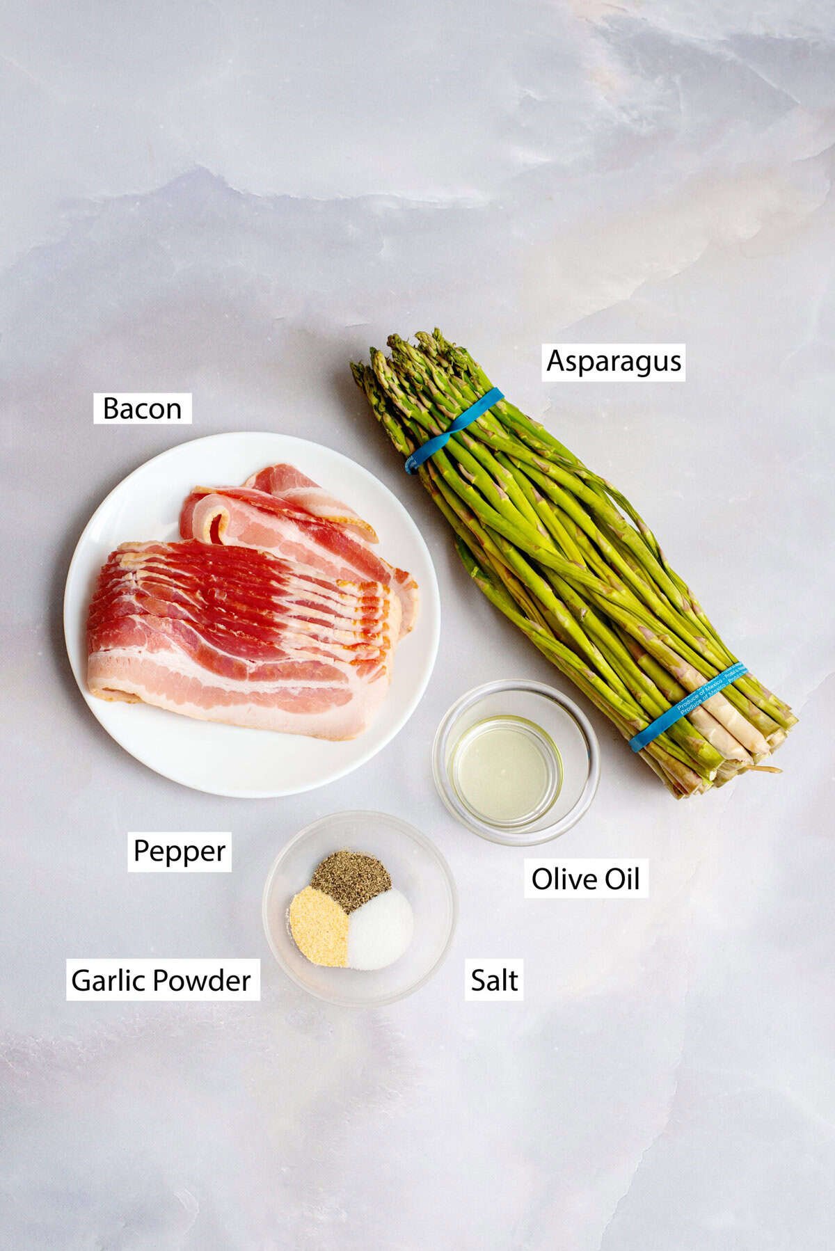 Ingredients for bacon-wrapped asparagus arranged on a light surface, including a bundle of fresh asparagus, a plate of raw bacon slices, a small bowl of olive oil, and a small dish with garlic powder, pepper, and salt. Each ingredient is labeled.