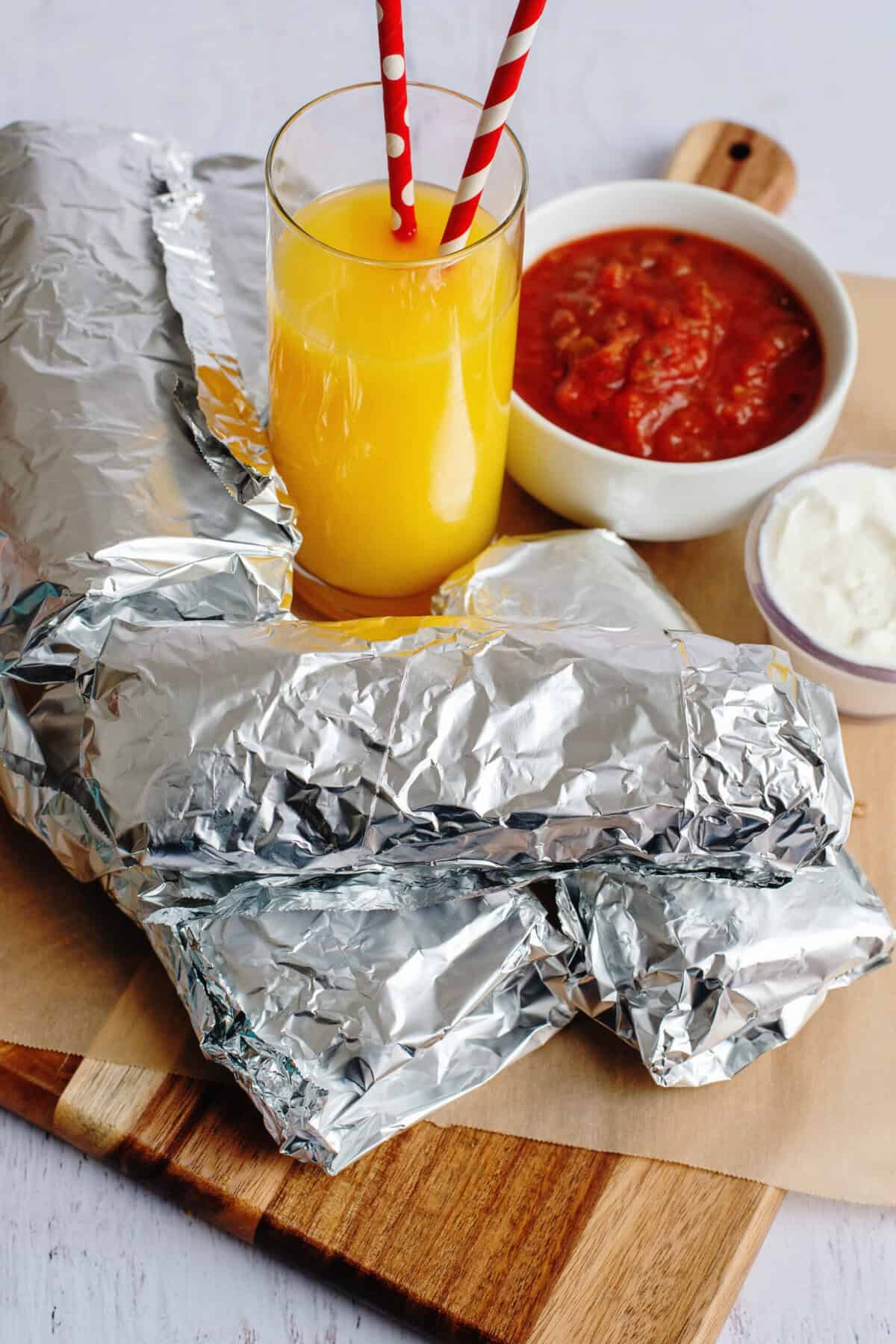 Grab and go breakfast burritos wrapped in foil on a tray with orange juice, salsa, and yogurt
