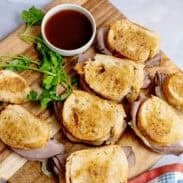 A cutting board with French Dips and au jus sauce