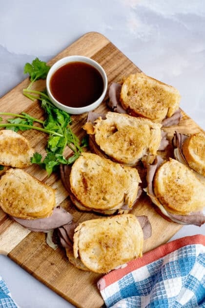 A cutting board with French Dips and au jus sauce