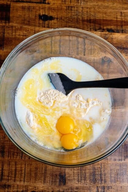 Stirring milk, butter, and eggs in a glass mixing bowl with a black rubber spatula.