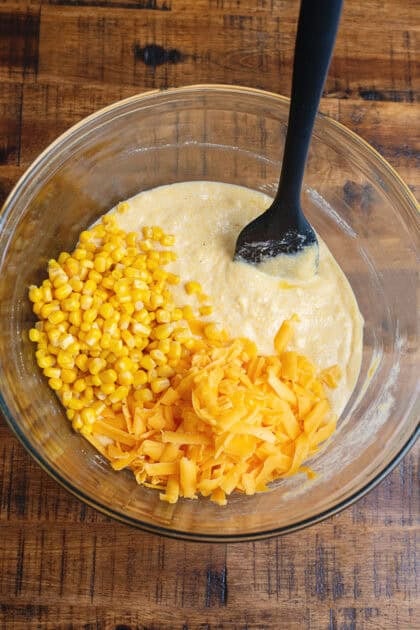 Stirring in cheese and corn into a glass mixing bowl with a black rubber spatula.