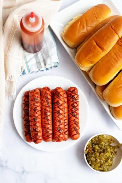 Grilled hot dogs on a plate next to condiments and buns