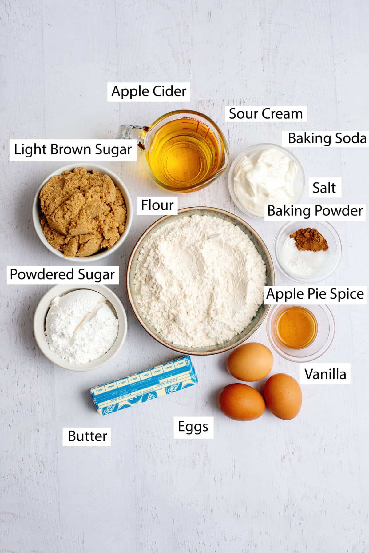 Ingredients for apple cider bundt cake, featuring all-purpose flour, baking powder, baking soda, salt, apple pie spice, butter, light brown sugar, eggs, apple cider, sour cream, vanilla extract, and powdered sugar.